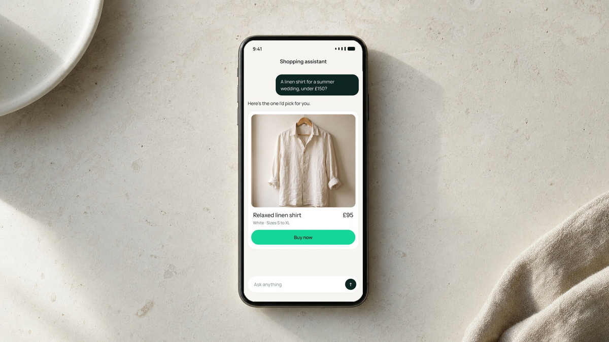 Phone on a stone table showing a shopper buying a linen shirt inside an AI chat, Shopify Editions Fall 2026